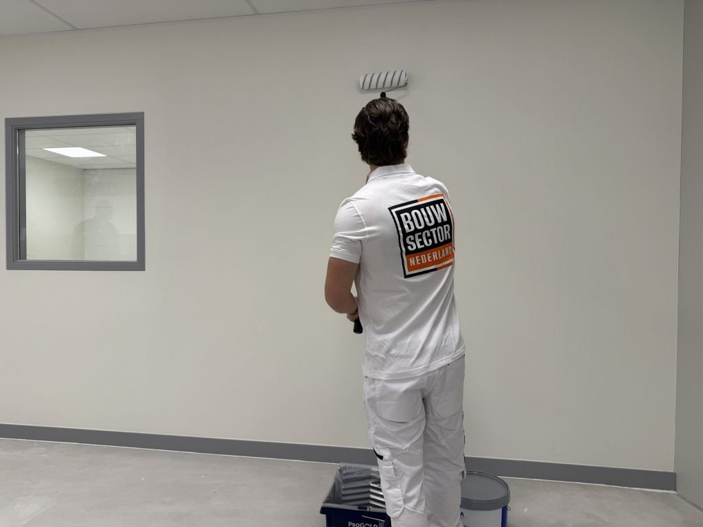 Schilder van Bouwsector Nederland rolt een lichte wand bij een binnenraam, met verfbak en emmer aan zijn voeten