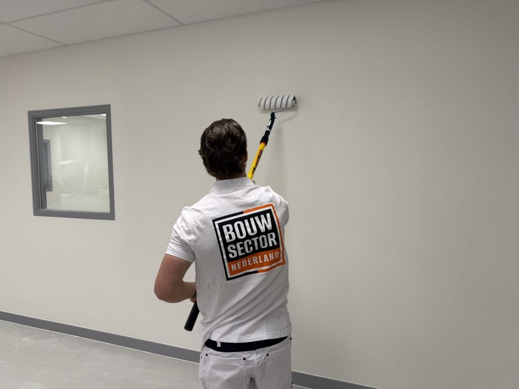 Schilder in wit werkpak zet de roller op een lichte wand naast een binnenraam