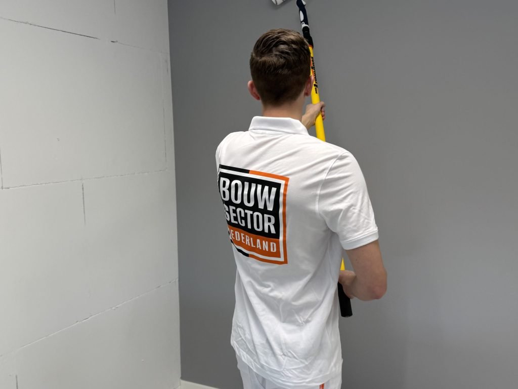 Schilder in wit poloshirt met logo van Bouwsector Nederland rolt een grijze wand naast een muur van kale blokken