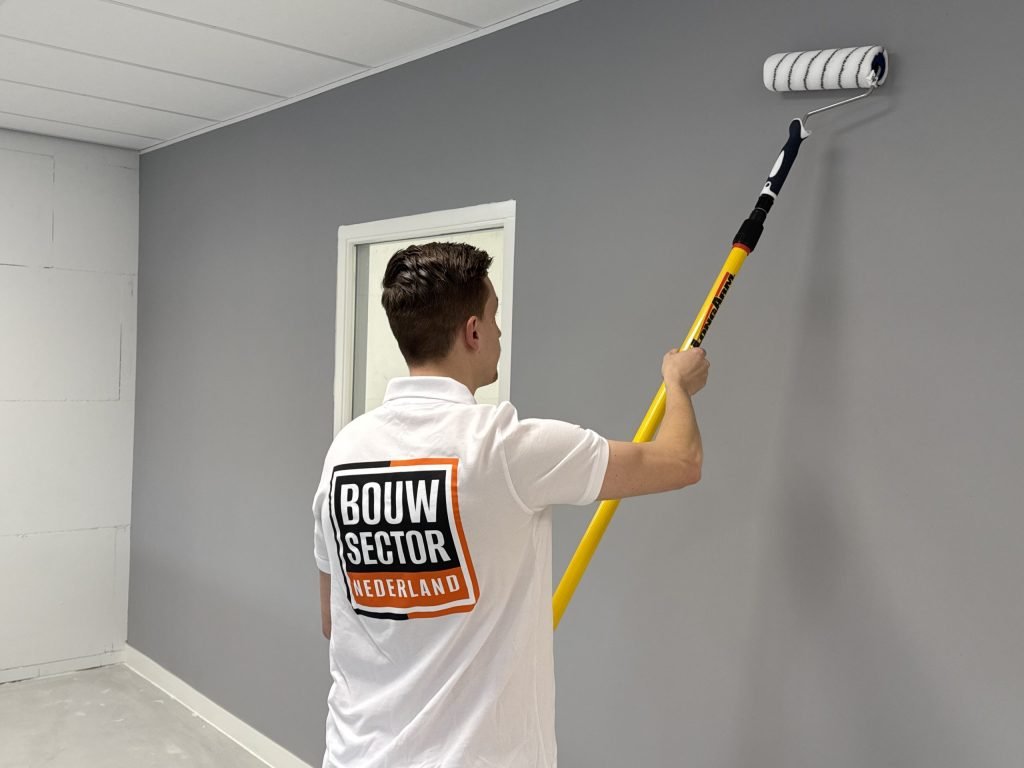 Grijze muur wordt tot tegen het plafond gesausd door een schilder met een lange verfroller