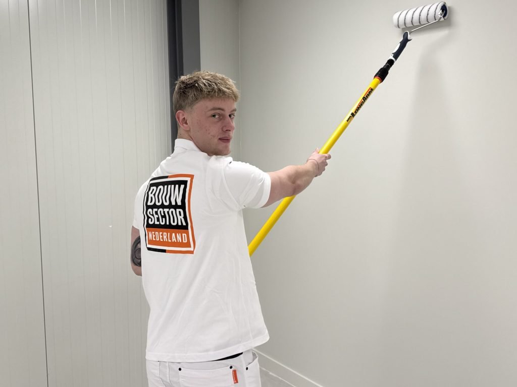 Jonge schilder van Bouwsector Nederland kijkt om terwijl hij met een verlengsteel een witte wand rolt