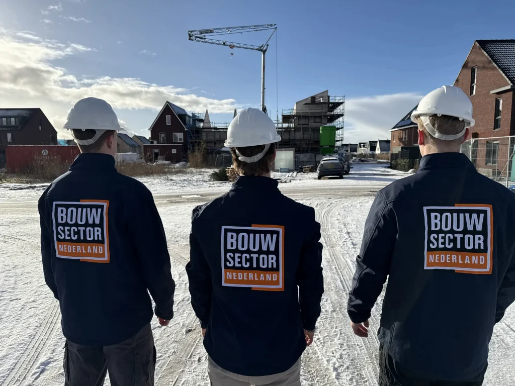 Drie vakmensen met witte helm en donkerblauwe jas van Bouwsector Nederland kijken naar een nieuwbouwwijk in de sneeuw
