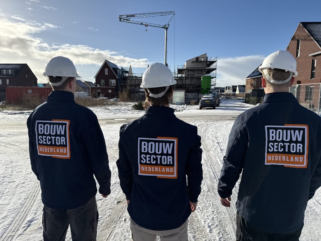 Drie medewerkers van Bouwsector Nederland met witte helm op de rug gezien bij nieuwe huizen in de steigers